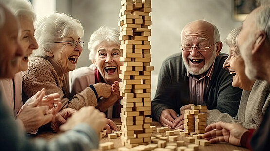 Das Bild zeigt ältere Menschen, die um einen Tisch sitzen. Sie lachen und spielen ein Spiel. In der Mitte des Tisches ist ein Turm zu sehen, den sie aus Holzklötzen bauen.