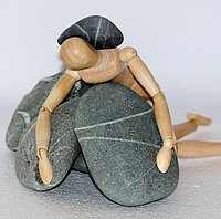 Ein Männchen aus Holz wird von dunklen Steinen niedergedrückt